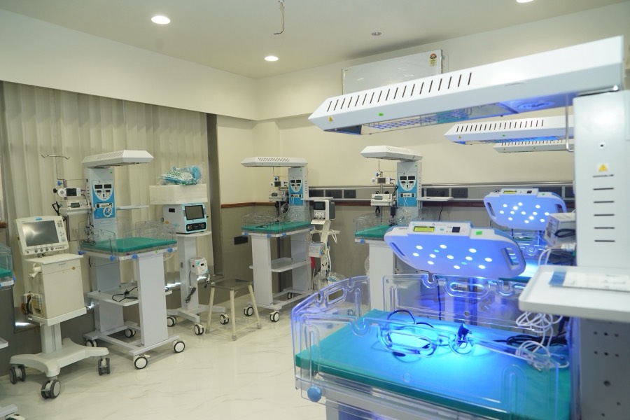 The neonatal intensive care unit: a row of radiant warmers with teal mattresses beside incubators glowing soft blue under phototherapy lights.