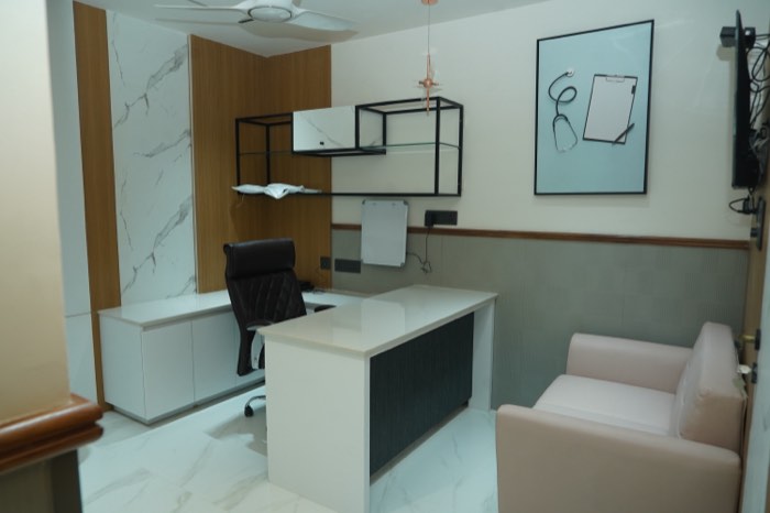 A third consulting room with a white desk, a leather chair, a pale sofa for visitors and framed stethoscope artwork on the wall.