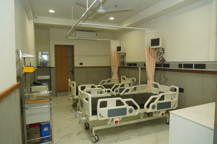 A multi-bed ward with adjustable beds, bedside monitors and blush privacy curtains on ceiling rails.