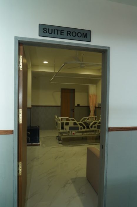 A doorway signed “Suite Room”, opening onto a made-up hospital bed and attendant seating.